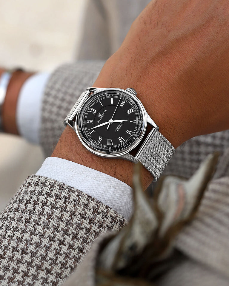 A round Automatic mens watch in Rhodiun plated 316L stainless from Waldor & Co. wit black double-layered dial in copper. Curved mineral glass with anti-reflective coating.Miyota 9093 movement. Mesh strap. The model is Continual 39 Amalfi.